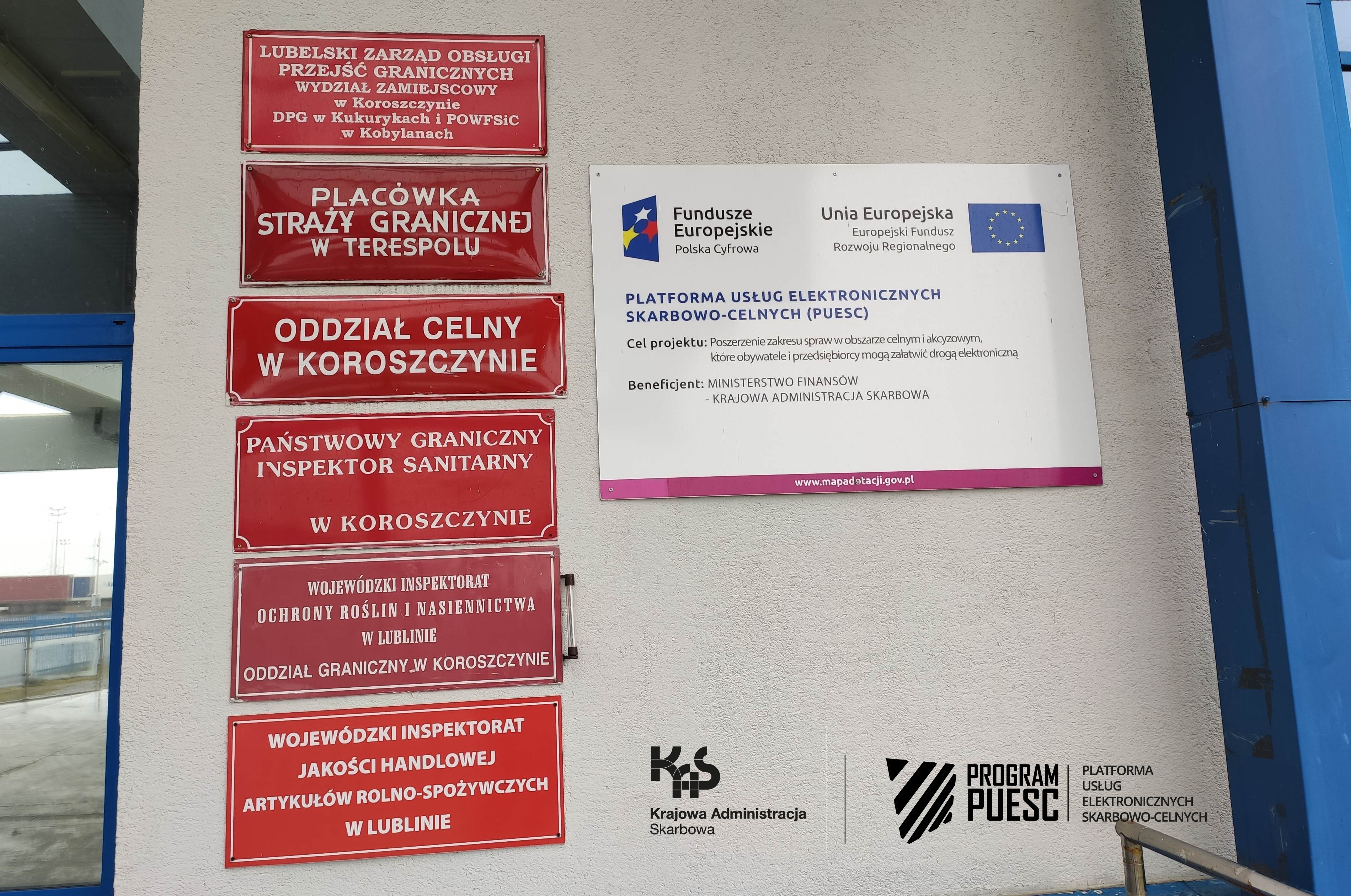 Na środku strony widać ścianę z czerwonymi tabliczkami informującymi o instytucjach mieszczących się w budynku. Na jednej z tablic umieszczona jest informacja o Projekcie PUESC i logotypy: Krajowa Administracja Skarbowa, Fundusze Europejskie Polska Cyfrowa, System Informacyjny Skarbowo-Celny, Ministerstwo Finansów, Unia Europejska Europejski Fundusz Rozwoju Regionalnego.