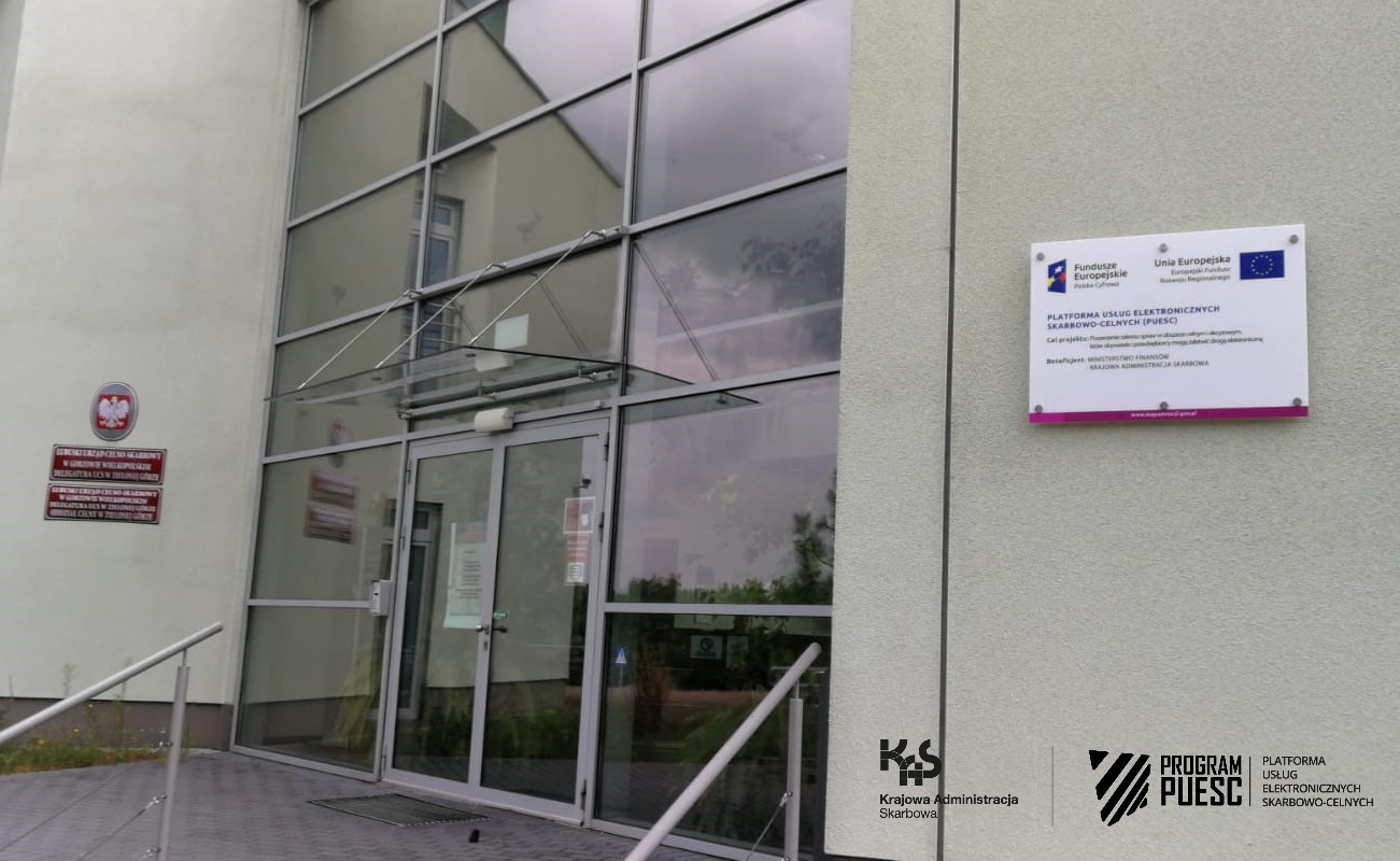 The main entrance to the building - Revenue Administration Chamber in Zielona Góra headquarters. In the center you can see a glass wall, on the left side there is the national emblem and red signs informing about the institutions located in the building. On the right, you can see a board informing about the implementation of the PUESC Project, with the following logos: National Revenue Administration, European Funds, Digital Poland, Tax and Customs Information System, Ministry of Finance, European Union, European Regional Development Fund.